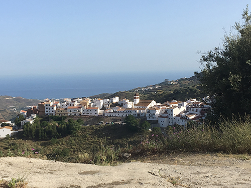 Un pueblo con casas blancas y tejados rojos ubicado en una colina con vegetación y el mar visible al fondo bajo un cielo despejado.

Descripción generada con IA