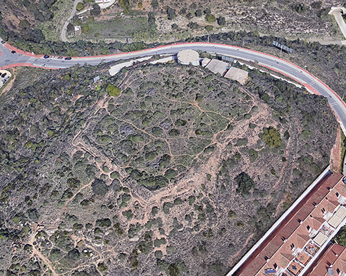 Terreno con vegetación baja y caminos de tierra rodeado por una carretera y edificios en un entorno urbano.

Descripción generada con IA